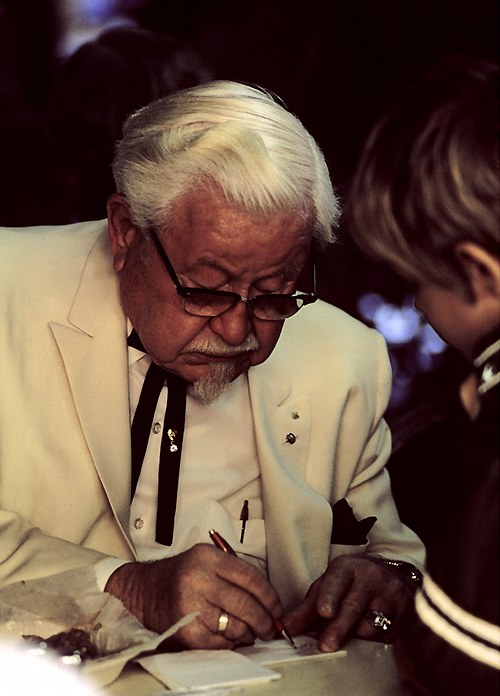 Colonel Harland Sanders in his white suit signing an autograph, around 1974