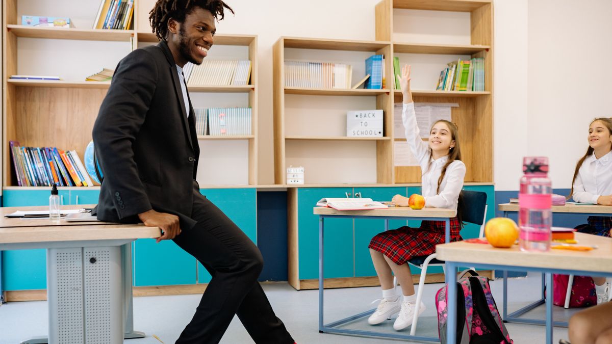 Teacher sitting on a desk while students raise their hands to answer a question