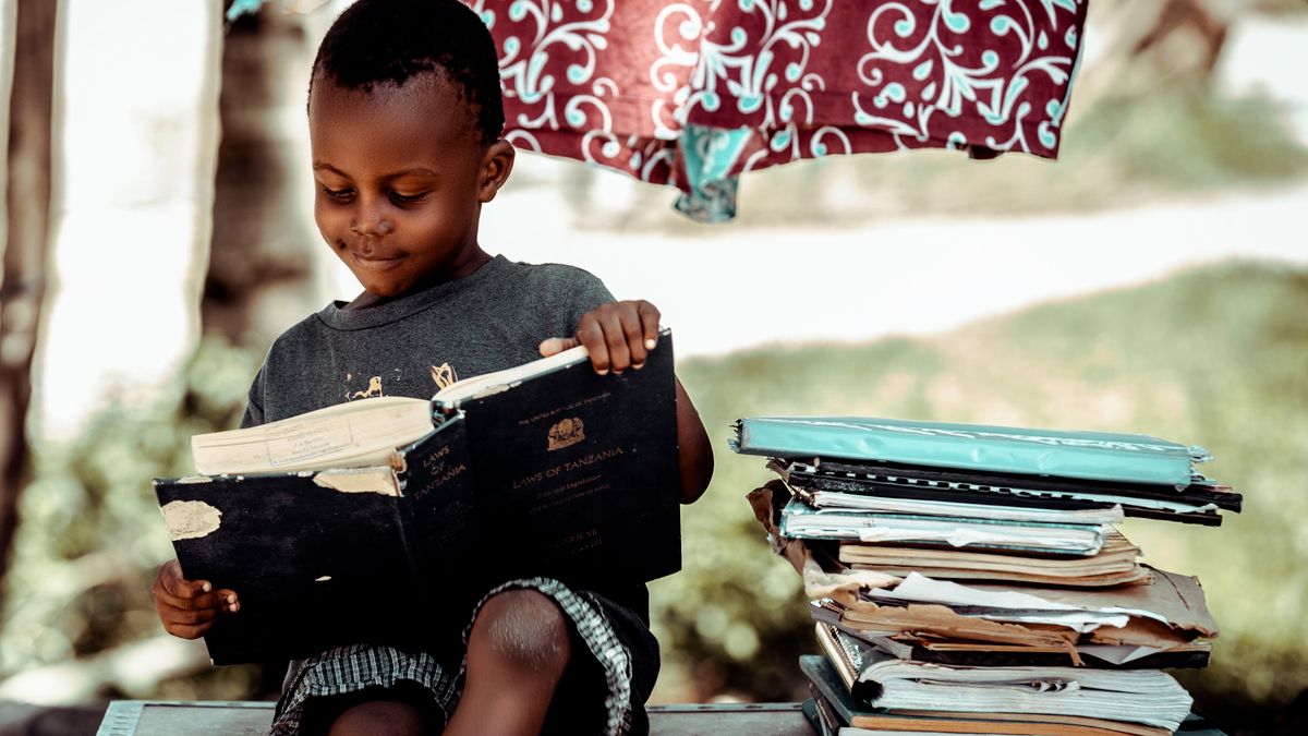 Mvulana mdogo wa Kitanzania akisoma kitabu nje, karibu na rundo la madaftari
